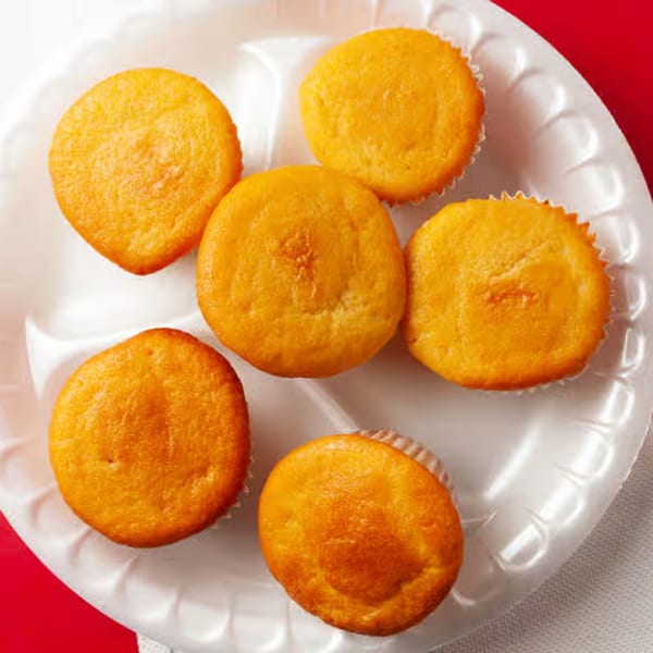 Six golden-brown corn muffins in white paper liners on a white disposable plate.